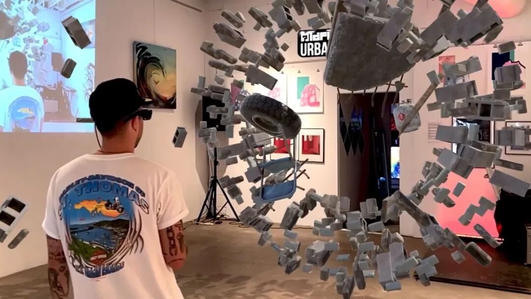 A person wearing a black cap and a white t-shirt with a surfing graphic stands in a gallery space, facing a circular installation made of metal frames holding multiple smartphone-like devices and screens displaying various images and videos. The setup includes floating elements suggesting augmented reality interaction, with art pieces on walls, a projector screen in the background showing similar devices, and additional props like tires integrated into the structure. The environment appears to be an indoor demo area for AR technology.